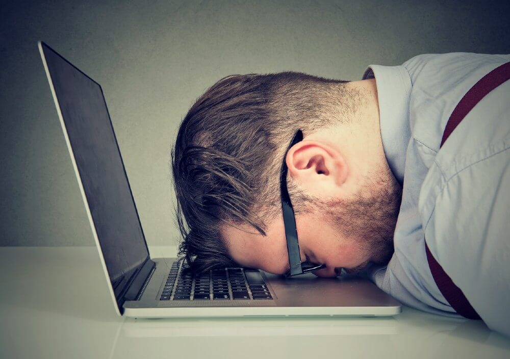 Frustrated man in glasses slumped over laptop, struggling with digital marketing tasks.