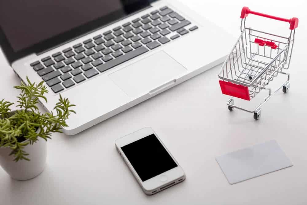 Laptop, smartphone, shopping cart, credit card, and plant on white desk for digital marketing and ecommerce concepts