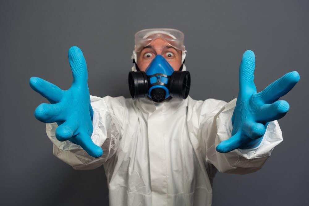 Person in white hazmat suit and blue gloves reaching forward, wearing a black respirator mask, gray background.