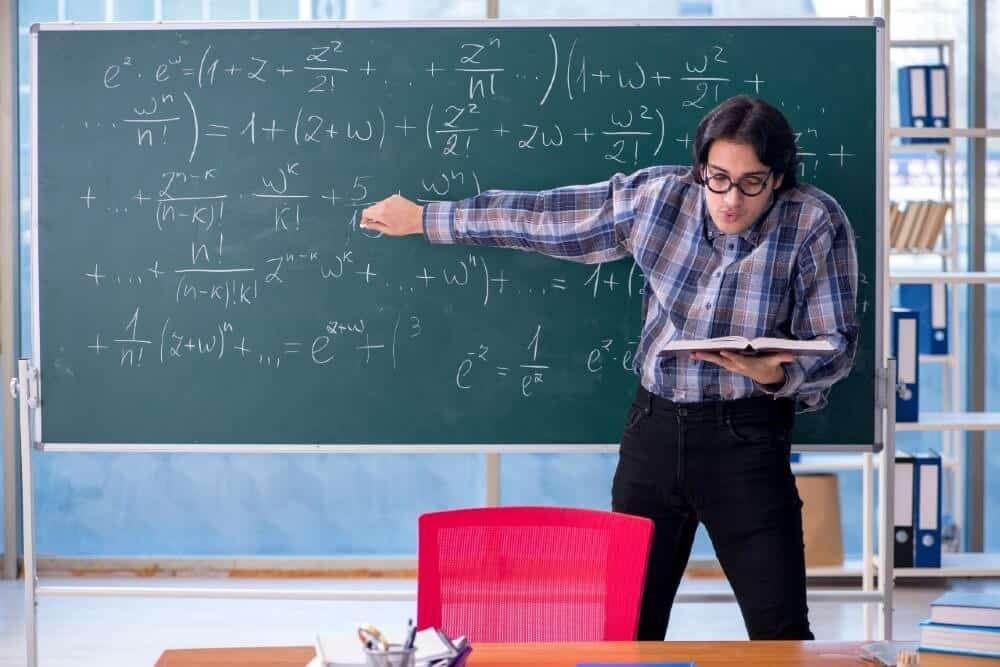 Teacher in a plaid shirt solving complex math equations on a green chalkboard in a classroom with red chair and shelves.