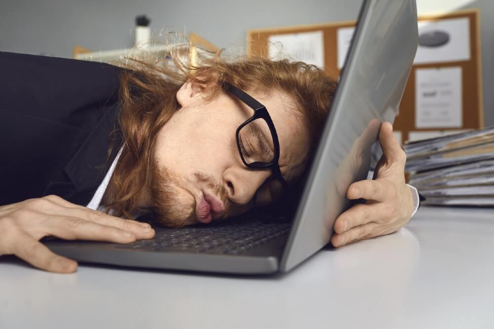 Exhausted worker with long hair asleep on gray laptop, cluttered desk, and office documents in digital marketing workspace.