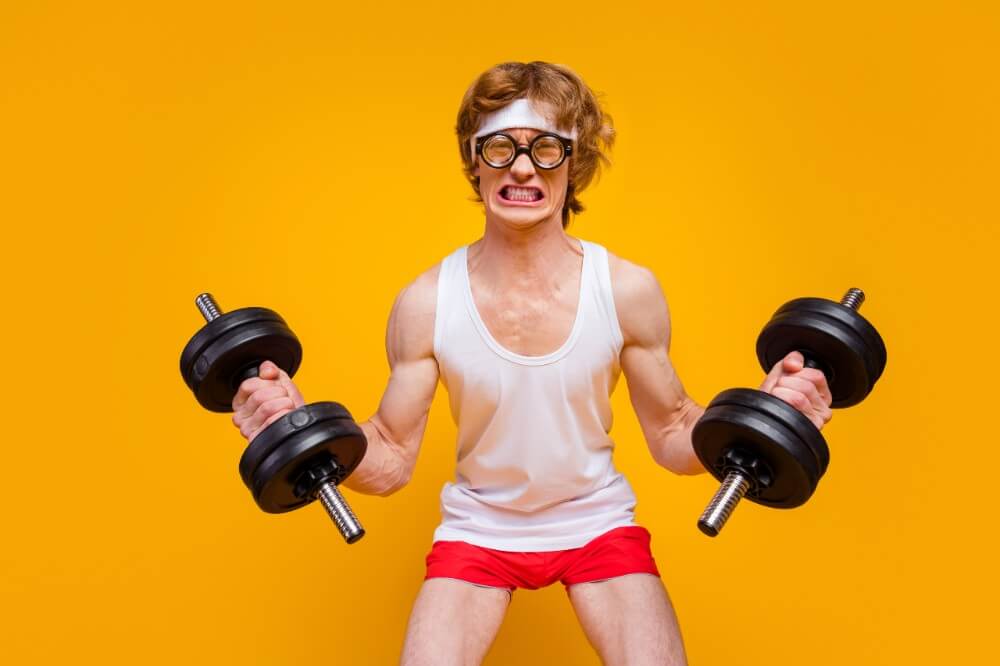 Man in red shorts and white tank top lifting dumbbells in front of bright yellow background, showcasing fitness effort and strength.
