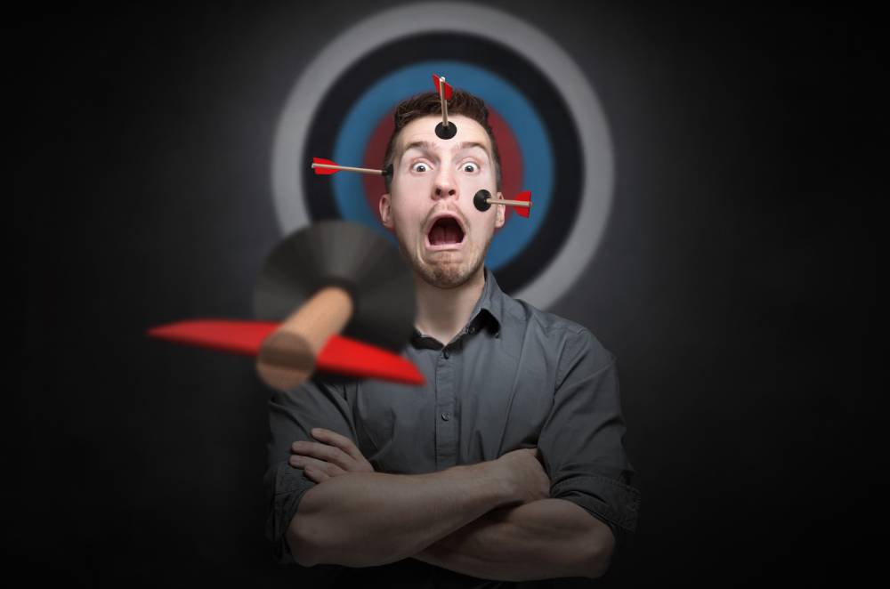 Man in gray shirt stands arms crossed near red bullseye target, arrow flying toward center on dark backdrop, digital marketing focus.